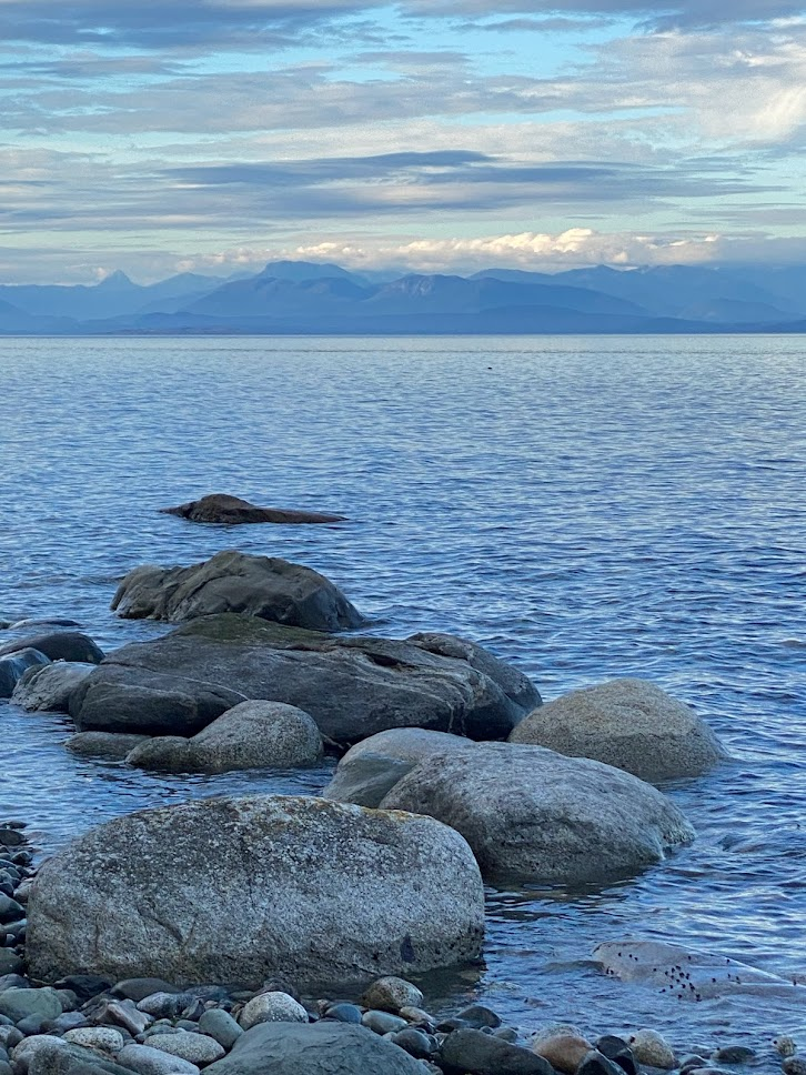 Powell River across the strait, taken by Andy Wynden A calm seascape with smooth mottled grey rocks in the foreground. Across the water there are mountains in the far distance. The photograph is taken in the early evening, and there are wisps of clouds all across the sky in various shades of blue and grey, complementing the blues and grays of the rocks and the ocean.